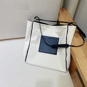 Un sac en tissu de luxe blanc posé sur une table blanche et adossée à un mur gris. À côté de lui, il y a une surface en bois clair. Ce sac a une bandoulière noire et une petite poche de couleur sombre à l'avant.