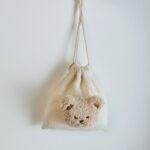 Un sac en toile beige avec un visage en peluche d'ourson. Il est suspendu à un mur blanc par ses hanses.