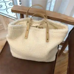 Un sac en peluche courte blanche posé sur une chaise marron. Celui-ci a deux hanses en simili cuir blanc. Derrière, il y a une baie vitrée et au sol du parquet en bois