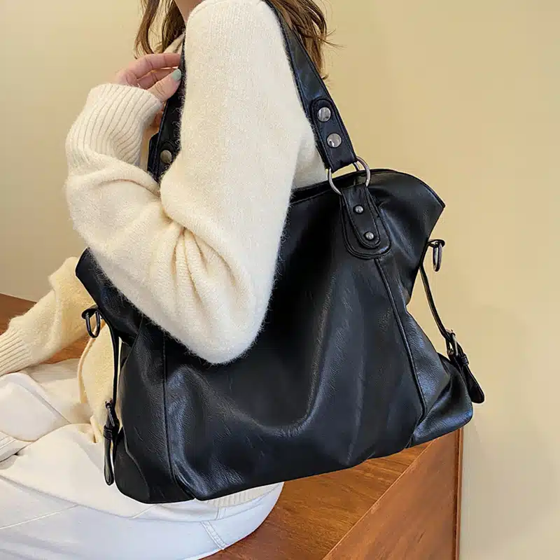 Une femme portant un grand sac à bandoulière en cuir Pu noir, elle le porte sur l'épaule et le tient avec sa main. Elle est habillée avec un pantalon blanc et un pull en laine beige. Elle est assise sur un meuble en bois marron.