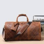 Un sac de voyage en cuir de style élégant et vintage avec une forme longue et arrondi. Il a deux hanses sur le dessus et une bandoulière qui tombe sur le côté. Il est posé sur une surface en bois de couleur marron.