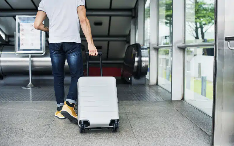 Un homme en train de marcher avec une valise à roulettes grise