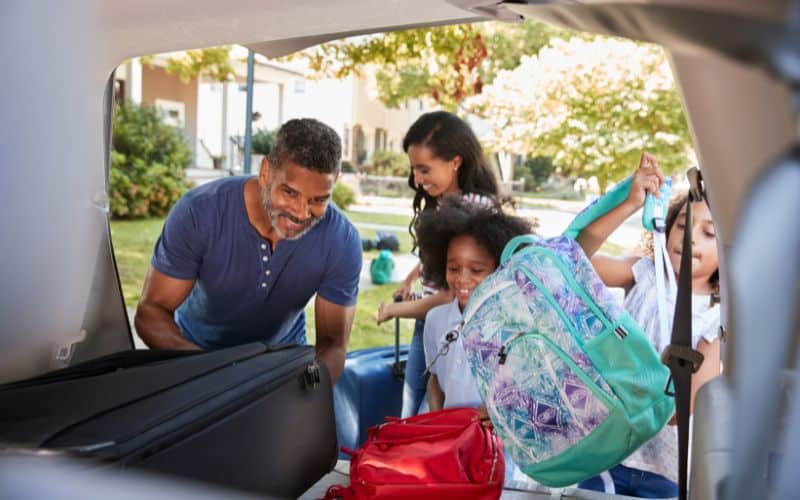 Famille composée de papa, maman et deux petites filles mettant leurs bagages dans le coffre d'une voiture pour les vacances.
