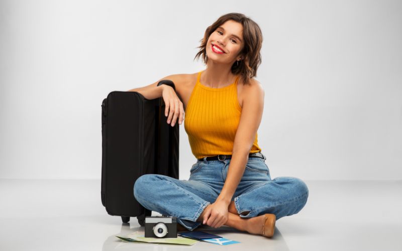 Femme assise en tailleur avec un grand sourire, à côté de sa petite valise-sac de voyage, un appareil photo est posé devant elle.