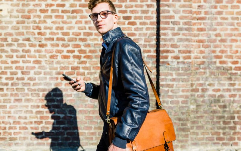 Homme portant une veste en cuir noire, des lunettes, un smartphone à la main et un sac d'ordinateur en cuir marron en bandoulière, il marche, un mur de briques derrière lui.