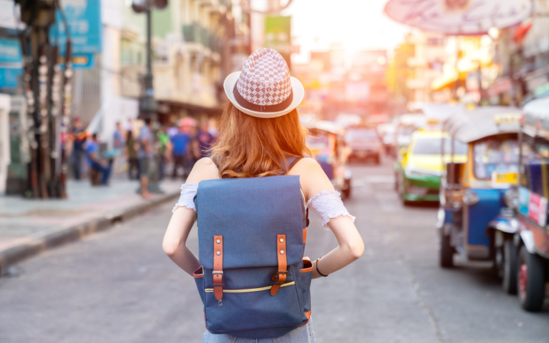 Jeune fille de dos, portant un chapeau, un sac à dos bleu avec des lanières en cuir. Dans une rue, on voit des gens sur le trottoir et des voitures.